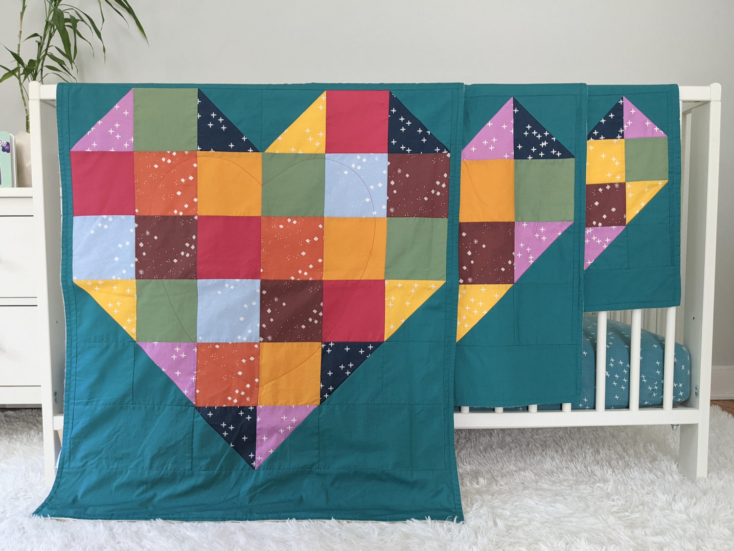 Baby Peipei Patchwork Heart. Three brightly colored, handmade, organic patchwork heart quilts hand on the side of a white crib. 