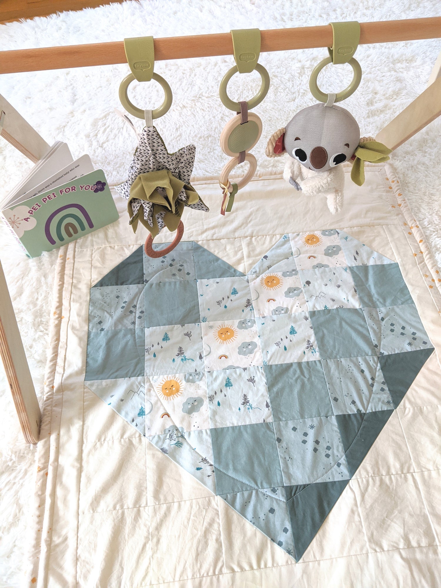 Baby Peipei, Ombre Heart in Green. A toy bar is positioned over a patchwork quilt whose blocks make up a heart. In the background is a book with text “A Peipei for You.” Tummy time and playtime are cozier with a peipei!