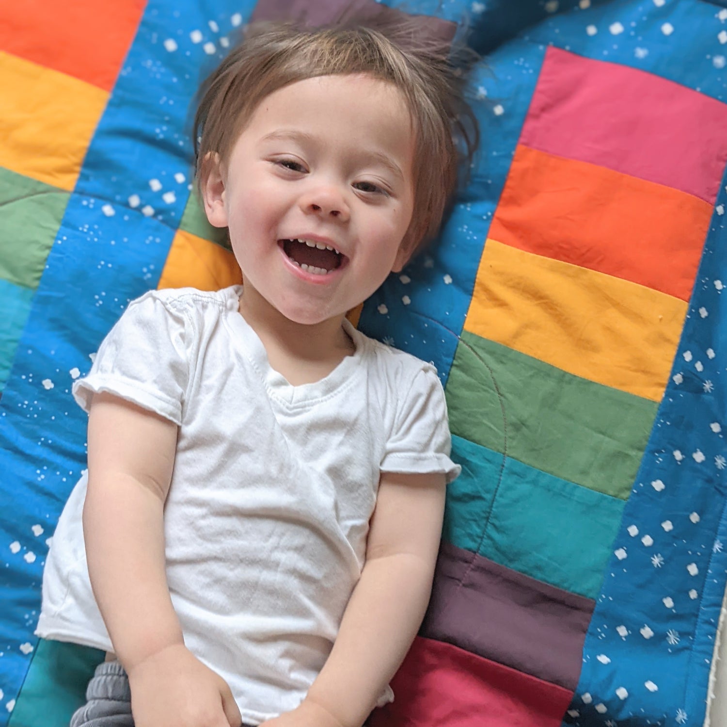 Baby Peipei, Bold Rainbow on Cloudy Blue background. Shot from above, a toddler smiles while lying on a quilt with rainbow stripes