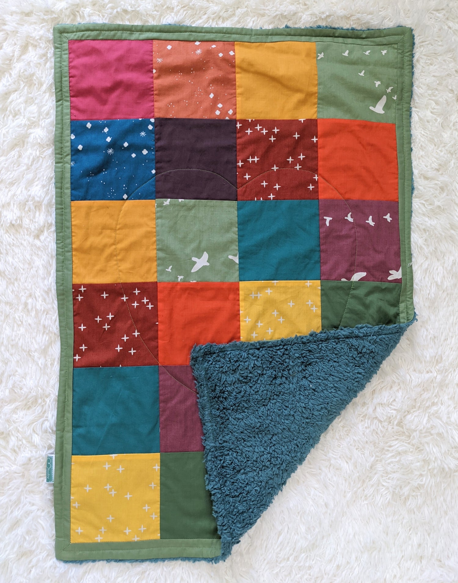 Baby Peipei, Rainbow Patchwork Midi. A multicolored, patchwork quilt lays on a white background. It measures 20 inches by 30 inches. The bottom right corner is folded showing the blue organic cotton Sherpa backing