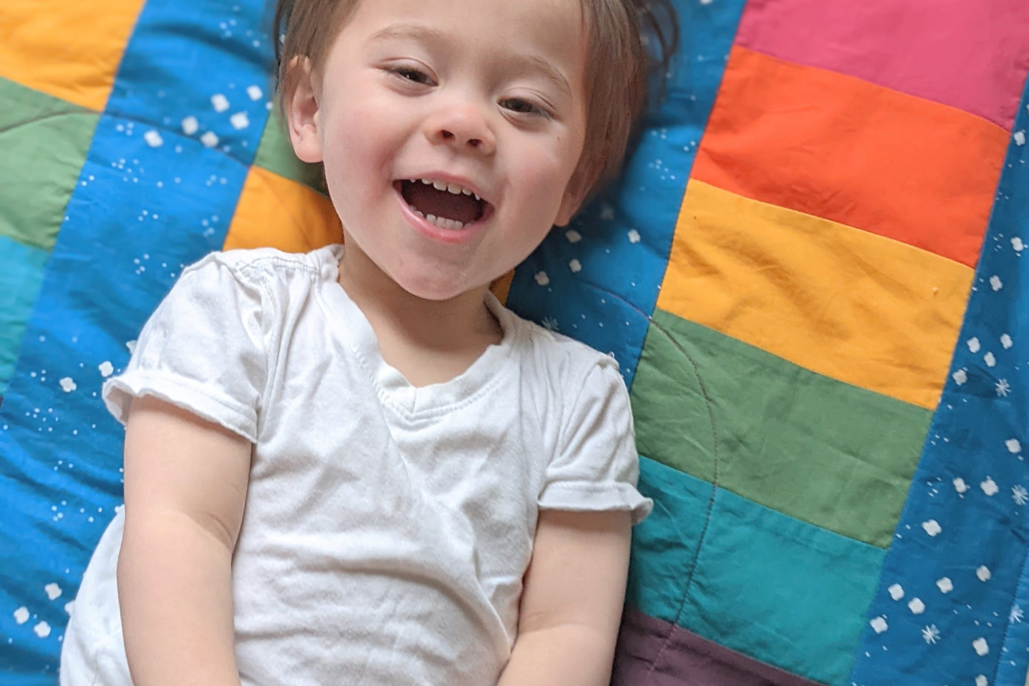 Baby Peipei, Bold Rainbow on Cloudy Blue background. Shot from above, a toddler smiles lying on a handmade organic quilt with rainbow stripes
